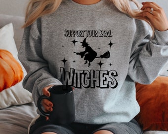 Op de afbeelding: Een grijs sweatshirt met een zwarte afbeelding van een heks die op een bezem vliegt met sterren en de tekst "Support Your Local Witches".