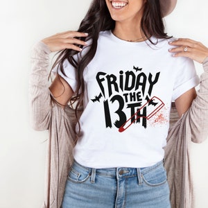 Könnte beinhalten: Weißes T-Shirt mit einer schwarzen Grafik, auf der "Freitag der 13." mit einem blutigen Messer und Fledermäusen steht.