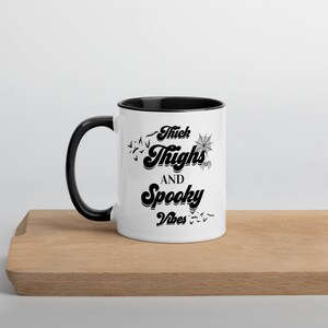 May include: A white ceramic mug with a black rim and handle. The mug features a black graphic that says "Thick Thighs and Spooky Vibes" with bats and a spider web.