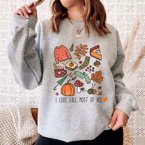 Könnte beinhalten: Grauer Pullover mit einem grafischen Druck von Herbstmotiven, darunter ein Kürbis, Blätter, eine Tasse heiße Schokolade, ein Pullover und ein Kuchen. Der Text "Ich liebe den Herbst am meisten" ist unter dem Grafikdruck aufgedruckt.