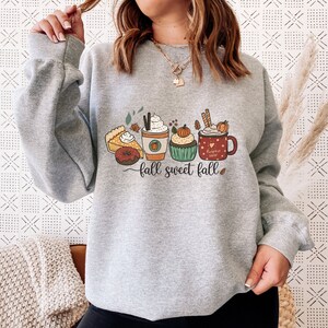 May include: Gray sweatshirt with a graphic of fall-themed treats, including a pumpkin spice latte, a cupcake, a slice of pie, a donut, and a mug that says "Pumpkin Spice Latte". The text "fall sweet fall" is printed below the graphic.
