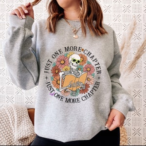 Op de afbeelding: Grijs sweatshirt met een afbeelding van een skelet met een zonnebril dat een boek leest, omringd door bloemen. De tekst "Just One More Chapter" wordt twee keer rond de afbeelding herhaald.
