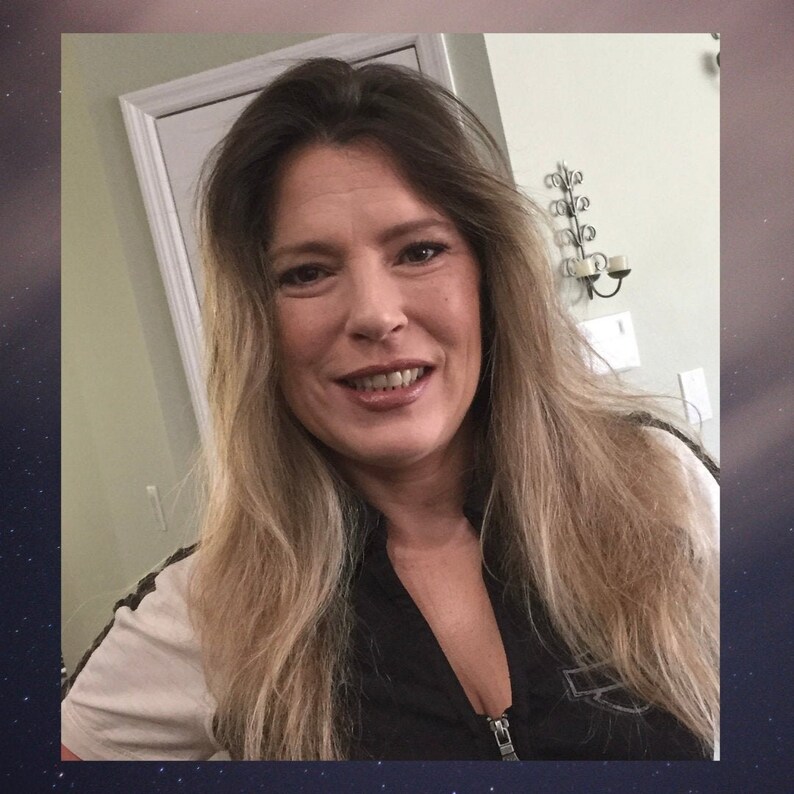 May include: A woman with long, wavy, light brown hair is wearing a black jacket with a zipper. She is smiling and looking at the camera.