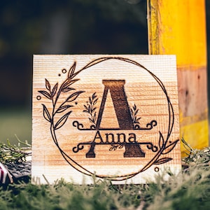 May include: A wooden sign with the letter "A" and the name "Anna" engraved in a circle with floral designs. The sign is on a green background.