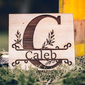 Puede incluir: Un letrero de madera con la letra "C" y el nombre "Caleb" grabados. El letrero está decorado con diseños florales.