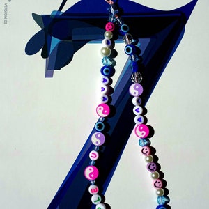 May include: A colourful beaded phone charm with a "PURPLE" word charm, yin-yang symbols, blue evil eyes, and smiley faces. The charm is hanging from a blue and white background.