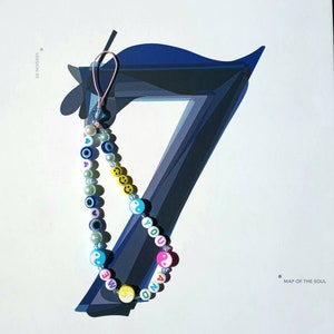 May include: A beaded phone charm with a pink, blue and yellow colour scheme. The beads spell out "YOU AND ME" with yin-yang symbols and smiley faces. The charm is hanging from a white background with the text "MAP OF THE SOUL 7 VERSION 02" printed on it.