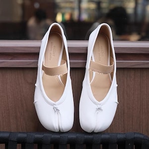 May include: A pair of white leather tabi shoes with a split toe design. The shoes have a simple, minimalist style and feature a beige elastic strap across the instep.