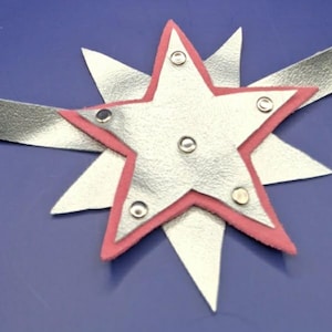 May include: A silver metallic headband featuring a layered star emblem. The emblem consists of a larger pink star base topped with a smaller silver star, secured by five silver-toned metal rivets. The band is a continuous strip of silver material.