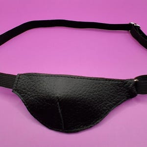 May include: A black eye patch featuring a textured leather-like surface with a contoured shape. The patch is attached to a thin, black elastic strap that includes a silver-toned metal adjustment buckle to secure the fit around the head.