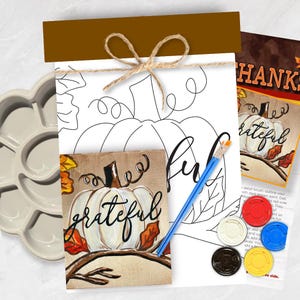 May include: A paint-by-number kit featuring a white pumpkin illustration with the word grateful, a brown header, twine bow, a blue paintbrush, a light gray paint palette, and five small circular paint pots in red, white, blue, yellow, and black.
