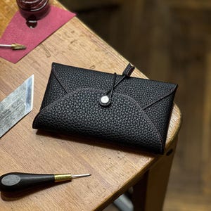 May include: A black leather wallet with a textured surface and a button closure. The wallet has a unique envelope-style design with red stitching. A small jar of red dye and a leather working tool are also visible.