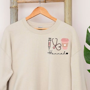 May include: A beige sweatshirt with a graphic design of a stethoscope, syringe, and coffee cup. The text "Hannah" is printed below the graphic.