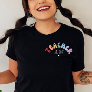May include: A black t-shirt with the text "TEACHER EST. 2023" in a rainbow arch design. The word "TEACHER" is in colorful bubble letters and the text "EST. 2023" is in white with a small red heart.