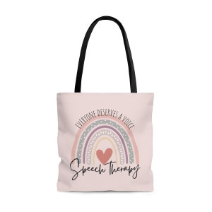 May include: A pink tote bag with black handles features a rainbow with a heart in the center. The rainbow has a gray, purple, pink, and yellow design with white polka dots. The text "Everyone deserves a voice" is written in a circle around the rainbow. The text "Speech Therapy" is written below the rainbow.