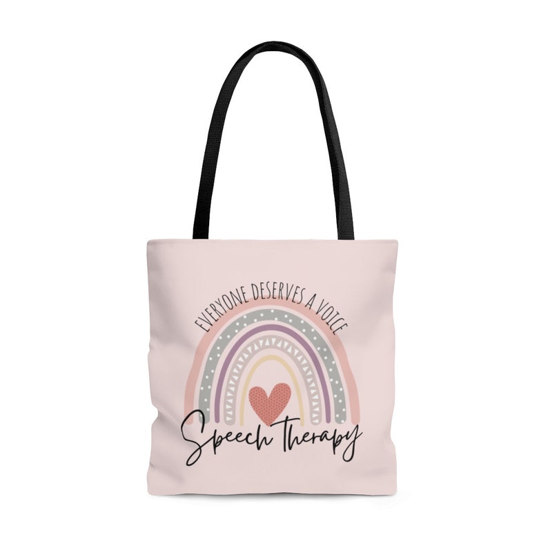 May include: A pink tote bag with black handles features a rainbow with a heart in the center. The rainbow has a gray, purple, pink, and yellow design with white polka dots. The text "Everyone deserves a voice" is written in a circle around the rainbow. The text "Speech Therapy" is written below the rainbow.