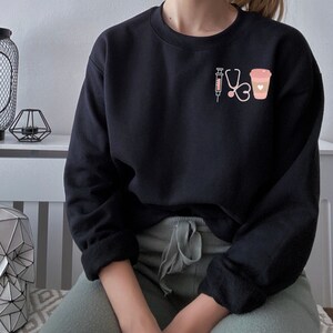May include: Black sweatshirt with a pink graphic of a syringe, stethoscope, heart, and coffee cup.