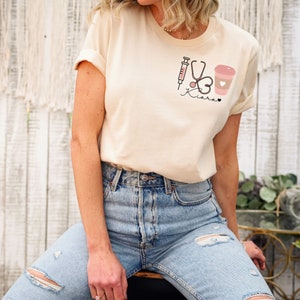 May include: A beige t-shirt with a pink syringe, stethoscope, heart, and coffee cup graphic. The text "Kiara" is written below the graphic.