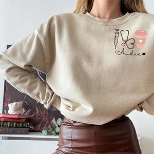 May include: A beige sweatshirt with a graphic design of a syringe, stethoscope, heart, and coffee cup. The text "Andie" is written below the graphic.
