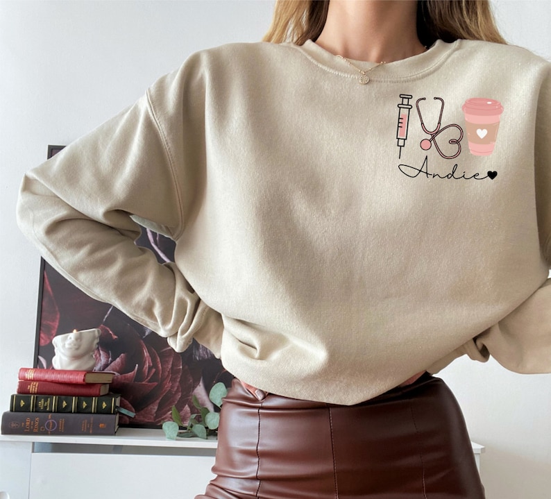 May include: A beige sweatshirt with a graphic design of a syringe, stethoscope, heart, and coffee cup. The text "Andie" is written below the graphic.