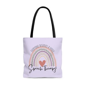 May include: A light purple tote bag with black handles. The bag features a rainbow with a heart in the center and the text "Everyone deserves a voice" above the rainbow and "Speech Therapy" below the rainbow.