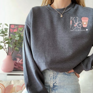 May include: A dark gray sweatshirt with a graphic design of a syringe, stethoscope, heart, and coffee cup. The text "Leslie" is written below the graphic.