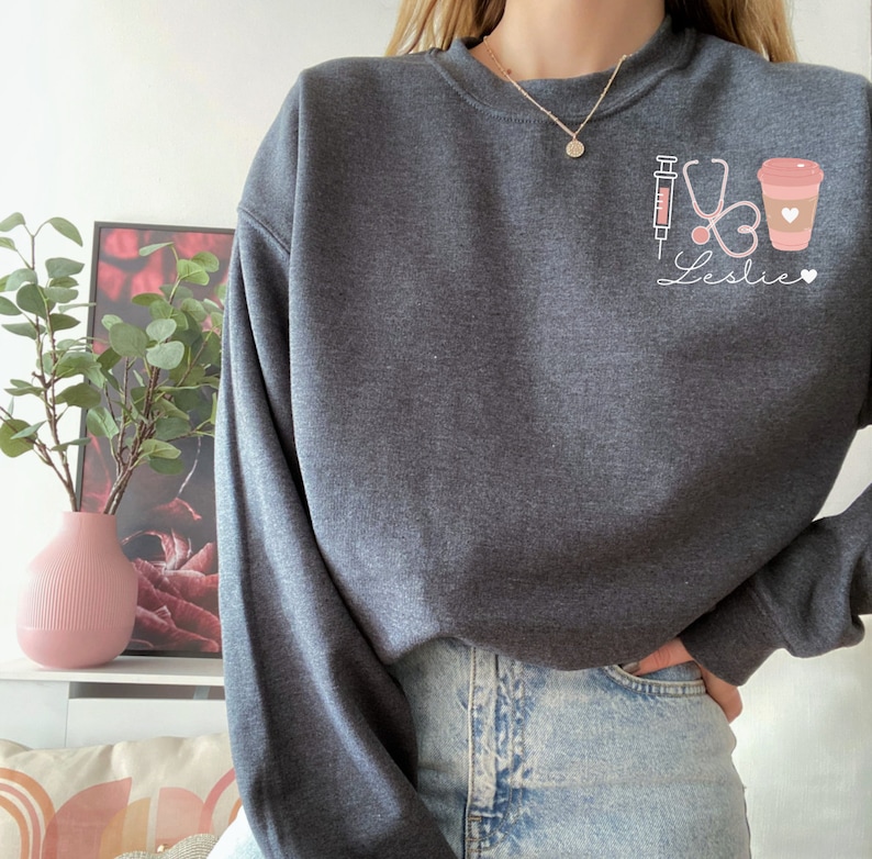 May include: A dark gray sweatshirt with a graphic design of a syringe, stethoscope, heart, and coffee cup. The text "Leslie" is written below the graphic.