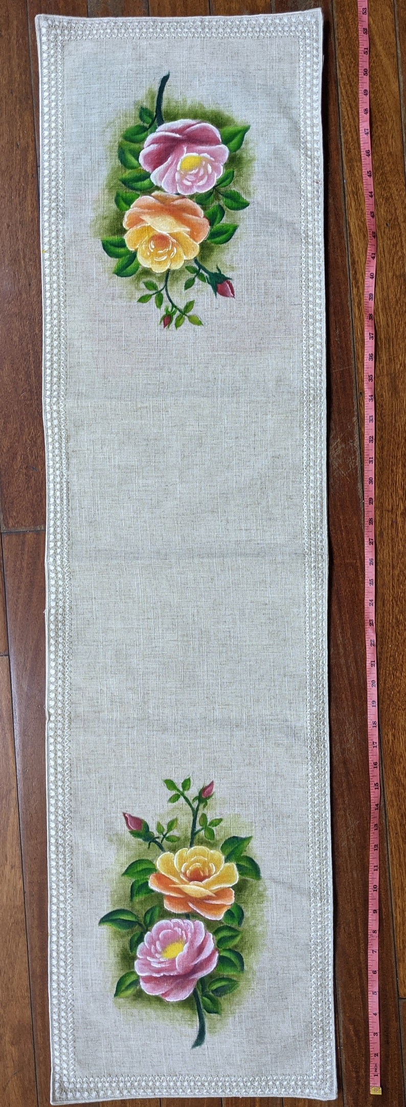 May include: Beige linen table runner with hand-painted roses in pink and yellow. The runner is approximately 48 inches long and features a decorative border. The roses are set against a green background.