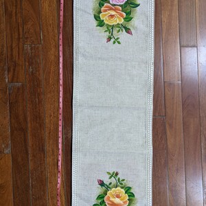 May include: A rectangular beige table runner with a floral design. The runner features hand-painted pink and yellow roses with green leaves. The runner is approximately 40 inches long and has a decorative border.
