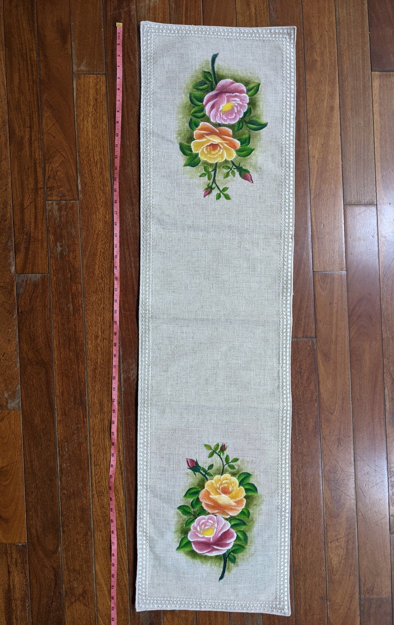 May include: A rectangular beige table runner with a floral design. The runner features hand-painted pink and yellow roses with green leaves. The runner is approximately 40 inches long and has a decorative border.
