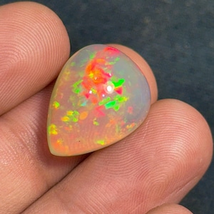 May include: A pear-shaped opal gemstone with a vibrant display of colors. The opal features flashes of red, orange, green, and yellow within a translucent, milky white base. The gemstone is held in a hand, showcasing its polished surface.