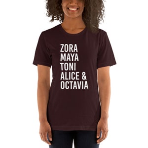 May include: A maroon t-shirt with white text that reads "Zora Maya Toni Alice & Octavia".
