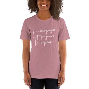 May include: A mauve-colored t-shirt with the French phrase "Le champagne est toujours la r&eacute;ponse" in white script. The shirt has short sleeves and a crew neck.