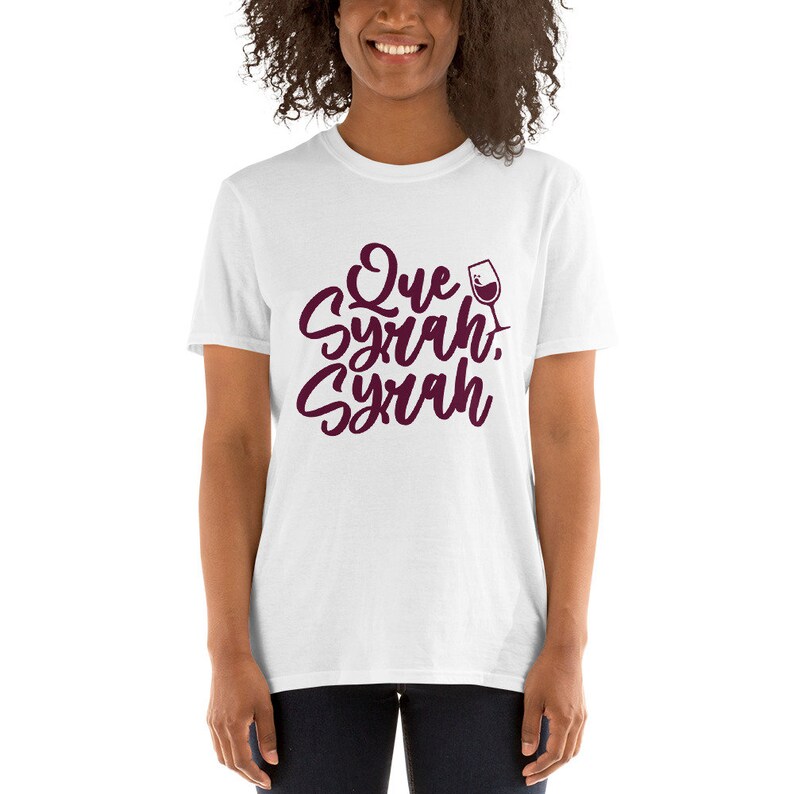 Puede incluir: Camiseta blanca con la frase "Que Syrah, Syrah" en escritura burdeos, acompa&ntilde;ada de un gr&aacute;fico de copa de vino. La camiseta es de cuello redondo y manga corta.