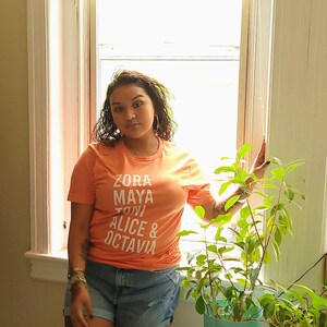 May include: A woman wearing a peach colored t-shirt with the names "Zora Maya Toni Alice & Octavia" printed in white. She is standing in front of a window with a plant in a pot.