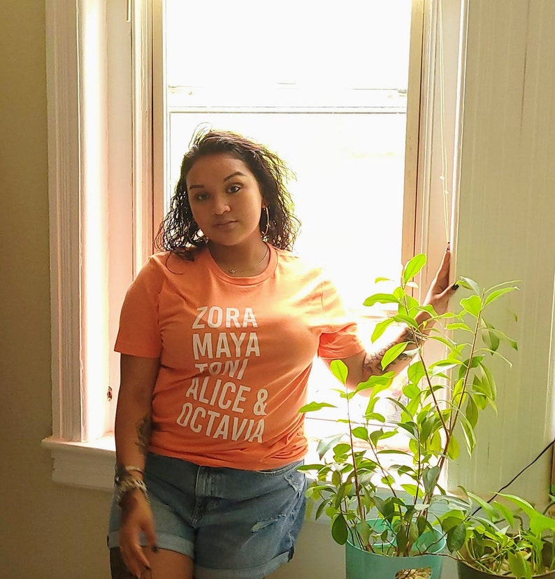 May include: A woman wearing a peach colored t-shirt with the names "Zora Maya Toni Alice & Octavia" printed in white. She is standing in front of a window with a plant in a pot.