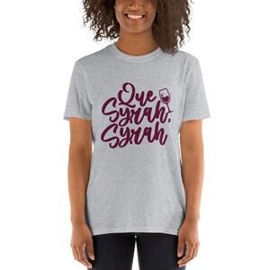 Puede incluir: Camiseta gris claro con la frase "Que Syrah, Syrah" en borgo&ntilde;a, en una fuente de escritura. Un gr&aacute;fico de copa de vino est&aacute; al lado del texto. La camiseta es de cuello redondo y manga corta.