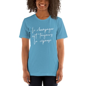 May include: A light blue t-shirt with the French text "Le champagne est toujours la r&eacute;ponse" in white script. The shirt is a crew neck style and appears to be made of soft material.