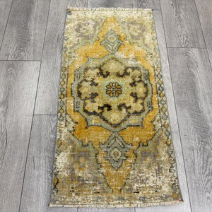 Puede incluir: Una alfombra rectangular de estilo vintage con un dise&ntilde;o de medall&oacute;n central. La alfombra presenta un aspecto desgastado con una paleta de colores amarillo, verde y marr&oacute;n. La alfombra est&aacute; colocada sobre un suelo de madera gris.