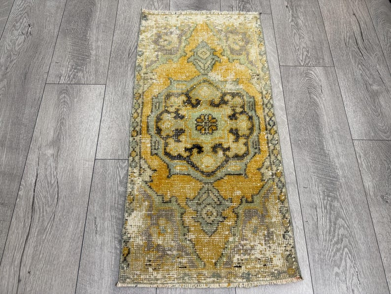 Puede incluir: Una alfombra rectangular de estilo vintage con un dise&ntilde;o de medall&oacute;n central. La alfombra presenta un aspecto desgastado con una paleta de colores amarillo, verde y marr&oacute;n. La alfombra est&aacute; colocada sobre un suelo de madera gris.