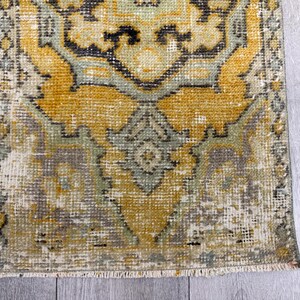 Puede incluir: Una alfombra rectangular desgastada con un patr&oacute;n tradicional. La alfombra presenta un dise&ntilde;o central en tonos de amarillo, gris y verde, con una apariencia descolorida. Los bordes tienen flecos y la alfombra est&aacute; colocada sobre una superficie de madera gris claro.