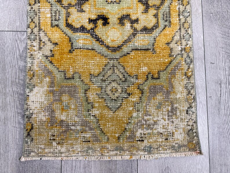 Puede incluir: Una alfombra rectangular desgastada con un patr&oacute;n tradicional. La alfombra presenta un dise&ntilde;o central en tonos de amarillo, gris y verde, con una apariencia descolorida. Los bordes tienen flecos y la alfombra est&aacute; colocada sobre una superficie de madera gris claro.