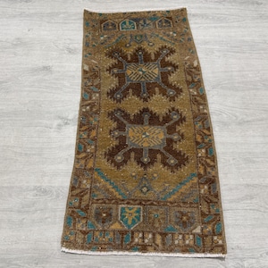 May include: A rectangular woven rug featuring a mustard yellow field with two prominent dark brown geometric medallions. The border displays intricate patterns in shades of brown, teal, and cream, with a short white fringe along the edges.
