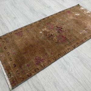 May include: A rectangular area rug featuring a distressed brown base with faded magenta and dark brown floral motifs. The rug has a repeating border pattern of small dark brown flower shapes and white fringe along the short edges.