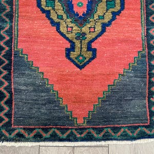 K&ouml;nnte beinhalten: Kleiner, rechteckiger Teppich mit einem korallenfarbenen Mittelfeld und einem dunkelgrauen geometrischen Design am unteren Rand. Der Teppich hat einen detaillierten Rand in Dunkelblau und Gr&uuml;n, mit einem zentralen Medaillon in Gold, Blau und Gr&uuml;n.