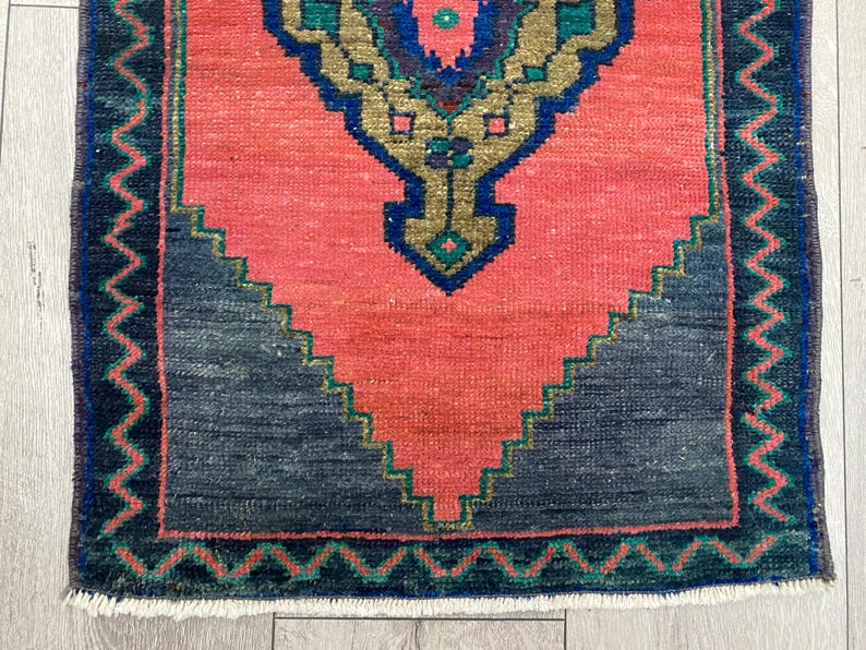 K&ouml;nnte beinhalten: Kleiner, rechteckiger Teppich mit einem korallenfarbenen Mittelfeld und einem dunkelgrauen geometrischen Design am unteren Rand. Der Teppich hat einen detaillierten Rand in Dunkelblau und Gr&uuml;n, mit einem zentralen Medaillon in Gold, Blau und Gr&uuml;n.