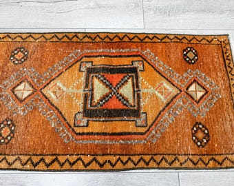 May include: A rectangular area rug featuring a vibrant burnt orange background with a central geometric diamond motif in black, cream, and muted green. The border displays a dark brown zigzag pattern, complemented by small circular floral accents.