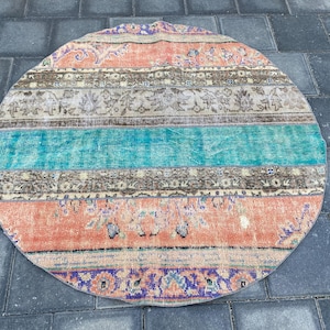 May include: Round, vintage rug with a patchwork design. The rug features a color palette of orange, teal, beige, and purple. The rug has a distressed look, with visible wear and tear, adding to its vintage charm. The rug is placed on a dark gray stone surface.
