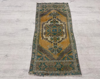 May include: A rectangular woven rug featuring a central diamond medallion in shades of mustard yellow, teal, and brown. The rug displays a geometric pattern with intricate borders and a fringed edge, showcasing a weathered, textured appearance.