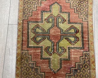 Puede incluir: Una alfombra rectangular con un motivo central de cruz geométrica en verde oliva y naranja óxido. El diseño incluye un borde de diamante escalonado y patrones de celosía complejos en las esquinas, en tonos marrón, verde y rojo apagado.